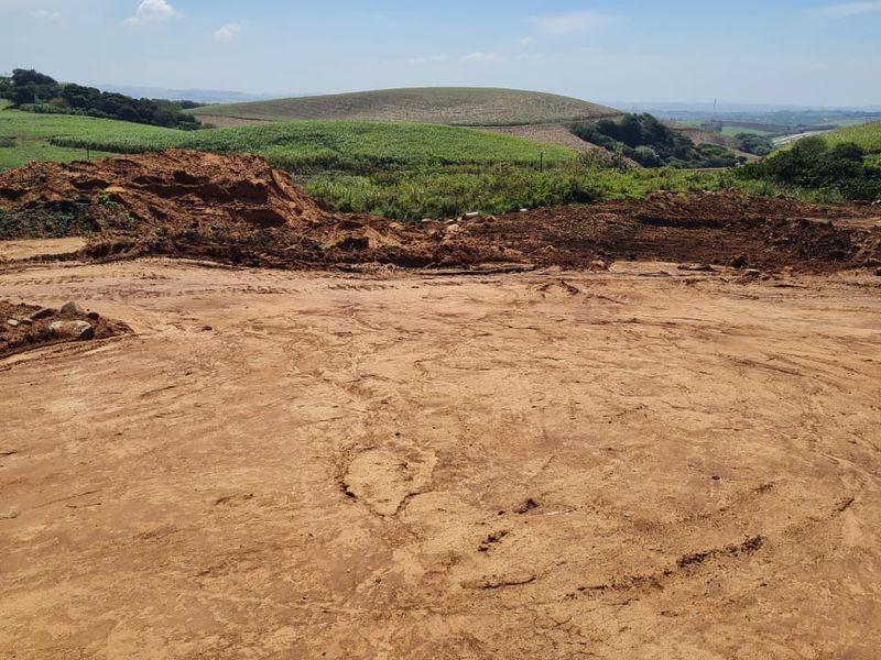 A view of a cleared residential plot featuring earthworks and surrounding rolling green hills under a clear blue sky.