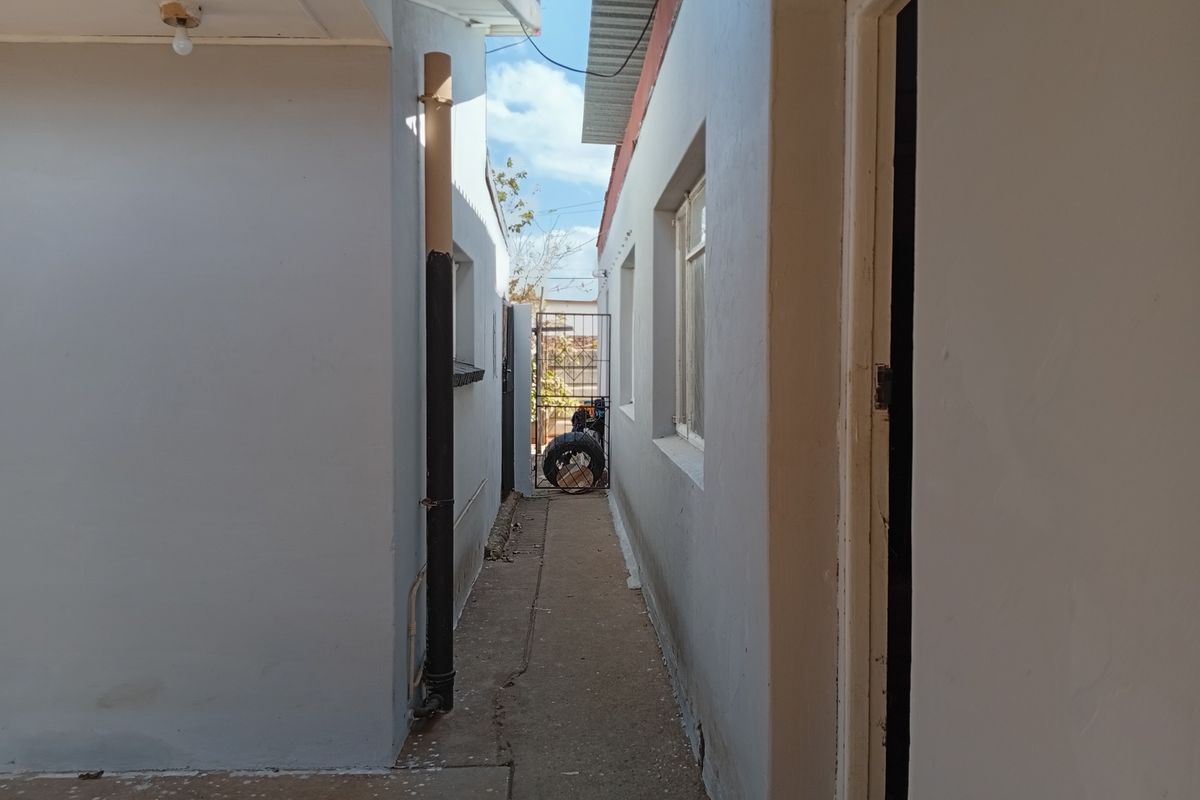 A narrow exterior side passage between two building structures, featuring a concrete pathway and metal security gate at the rear.