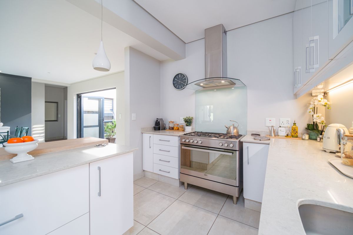 Modern kitchen featuring white cabinetry, stone countertops, stainless steel gas range with extractor hood, and tiled flooring, seamlessly flowing into the adjacent living area.