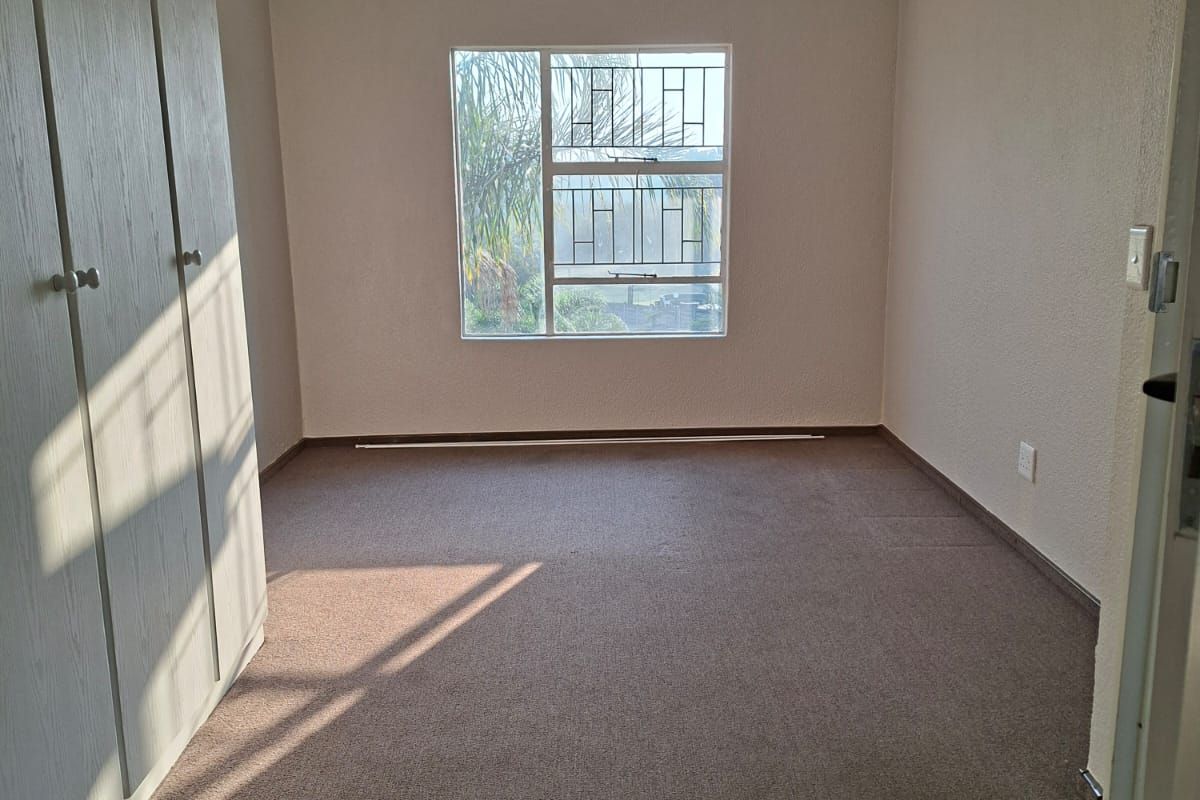 A clean, empty bedroom featuring built-in cupboards, carpeted flooring, and a window fitted with security bars.