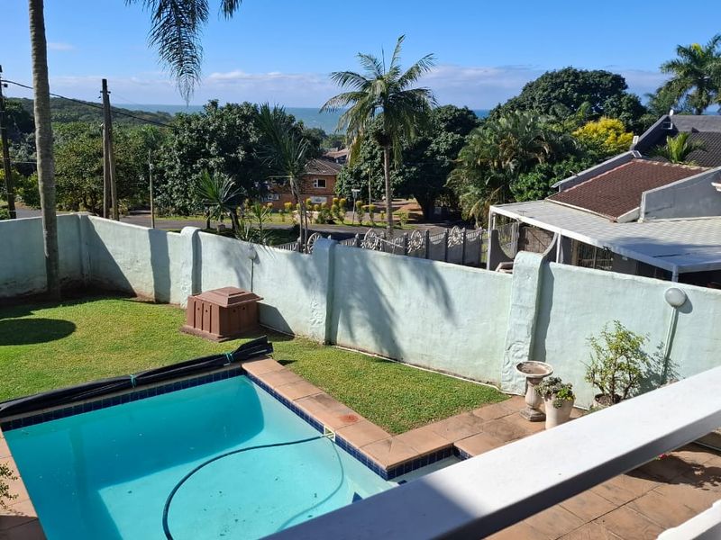 An elevated view of a private backyard featuring a swimming pool, a small lawn area, and a perimeter wall with distant ocean views.