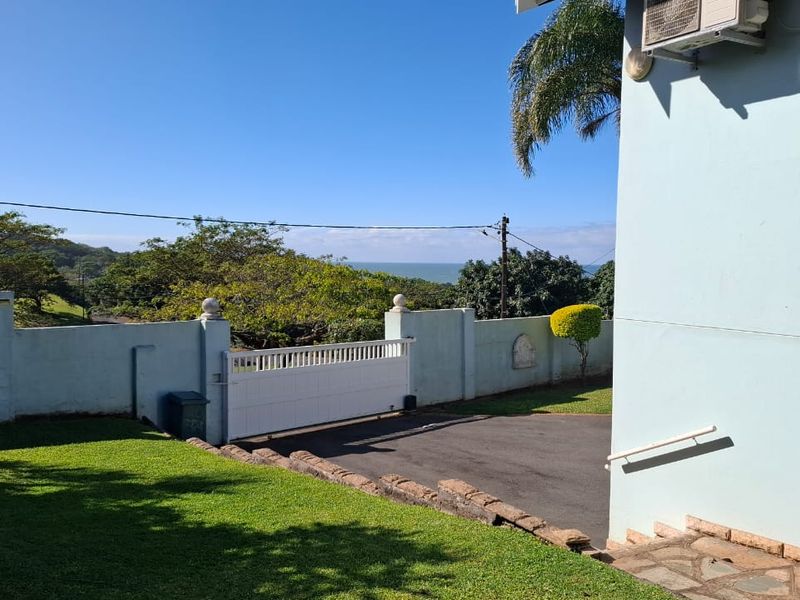 Front exterior view featuring a paved driveway, secure sliding gate, boundary wall, and distant ocean views.