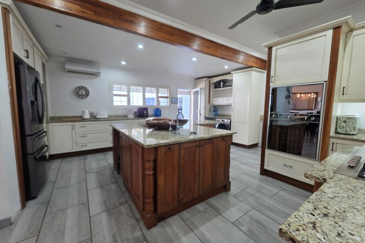 Spacious kitchen featuring a large central island with granite countertops, custom cabinetry, built-in oven, and modern tiled flooring under an exposed timber beam.