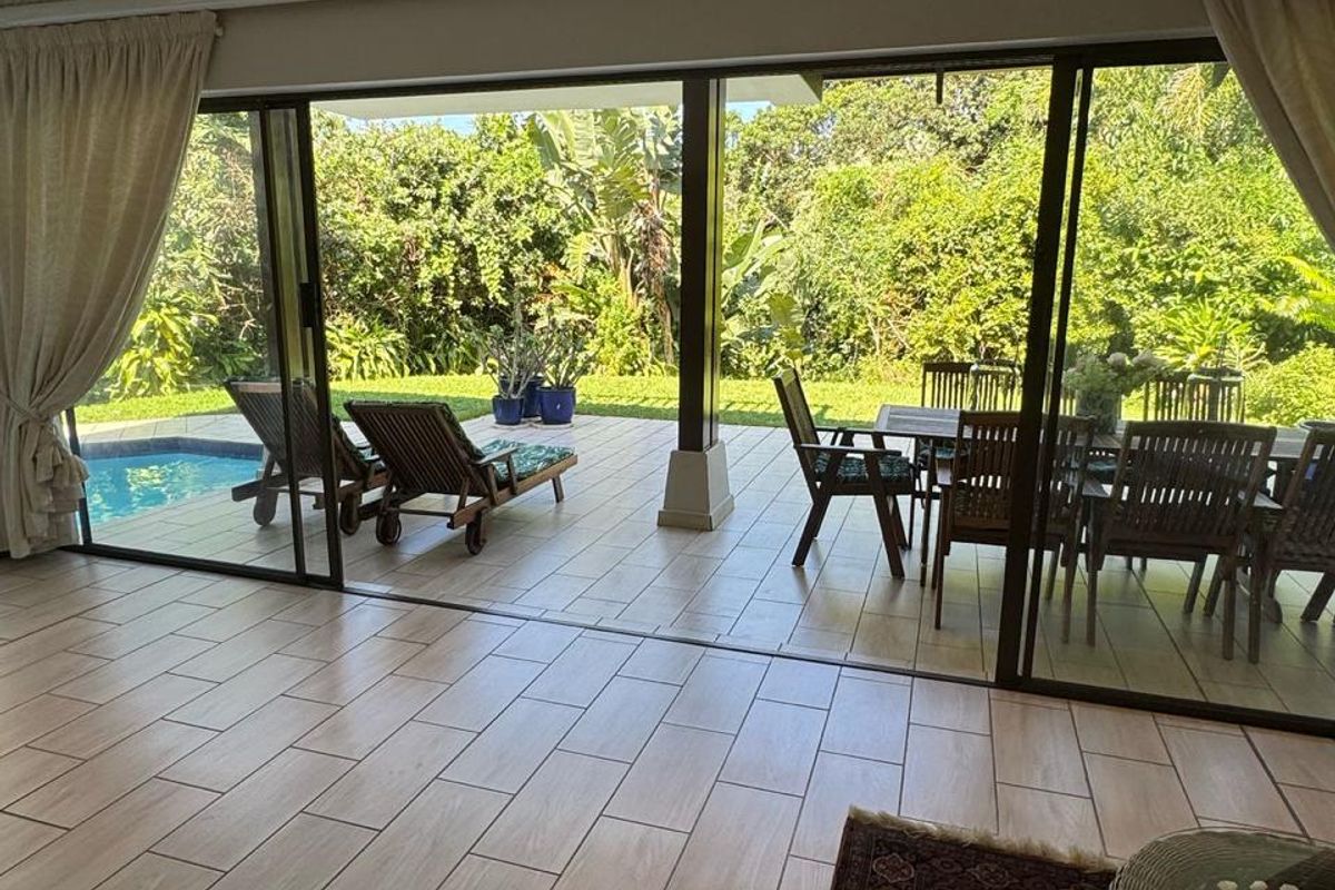 Spacious living area featuring large sliding glass doors that open onto a covered patio, swimming pool, and lush garden views.