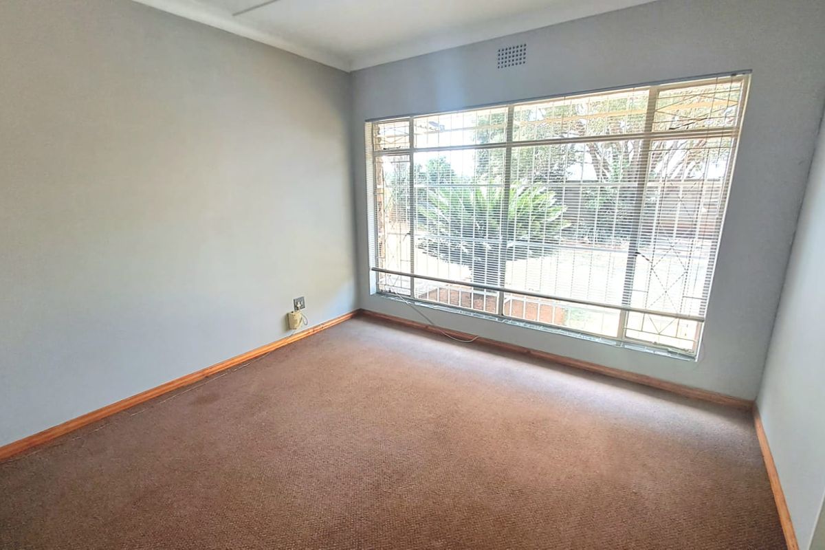 A vacant bedroom featuring neutral walls, brown carpeting, and a large window with security bars overlooking the garden.
