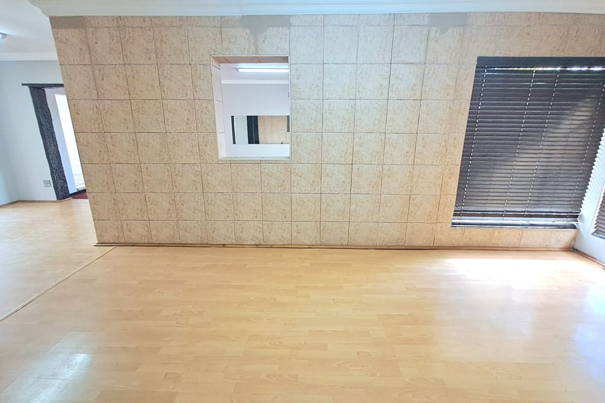 An empty living area featuring light-toned laminate flooring, a tiled feature wall with a service window, and dark horizontal window blinds.