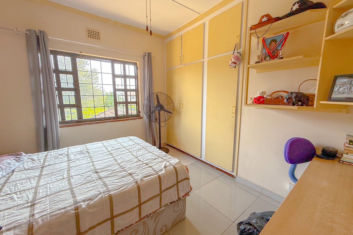 A bedroom featuring built-in cupboards, tiled flooring, and a large window with security bars, offering a functional and well-lit living space.