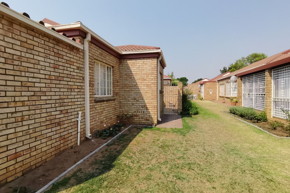 A view of the rear exterior of the townhouse, featuring face brick walls, tiled roofing, security bars on windows, and a well-maintained lawn area.