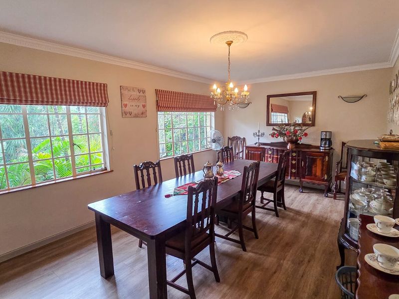 A spacious dining room featuring laminate flooring, crown molding, a decorative ceiling rose with a chandelier, and large windows overlooking the garden.