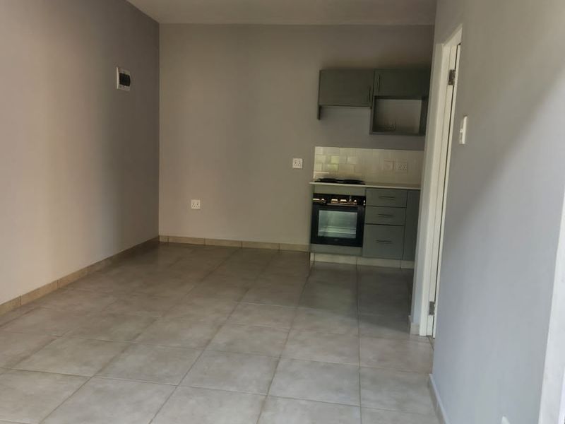 An open-plan living area featuring neutral tiled flooring and a compact, functional kitchen with built-in cabinetry, oven, and hob.