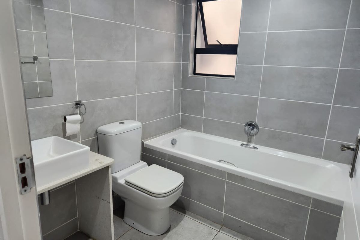 A modern, clean bathroom featuring grey wall and floor tiles, a built-in bathtub, a white toilet, and a wall-mounted basin.