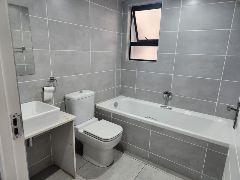 A modern, clean bathroom featuring grey wall and floor tiles, a built-in bathtub, a white toilet, and a wall-mounted basin.