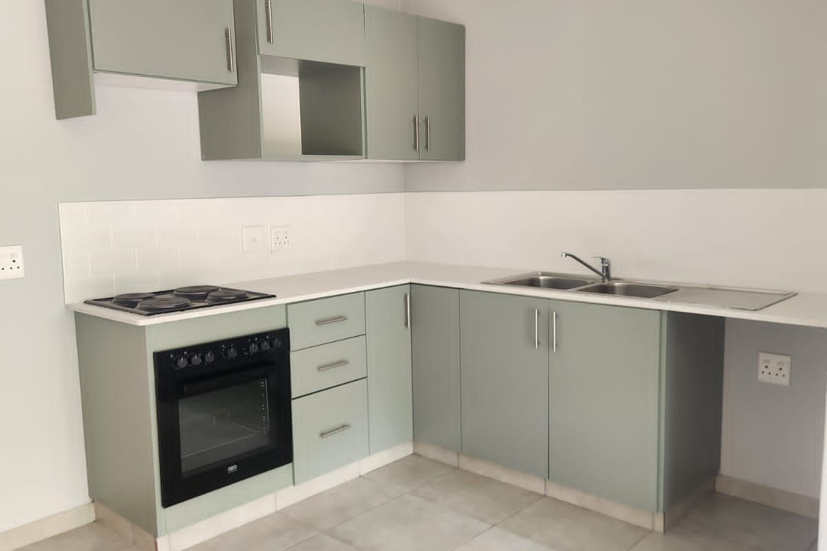 A clean, modern kitchen featuring sage green cabinetry, a built-in oven and hob, stainless steel sink, and neutral tiled flooring.