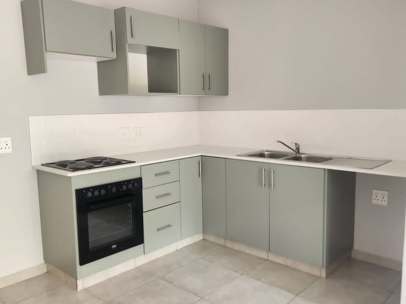 A clean, modern kitchen featuring sage green cabinetry, a built-in oven and hob, stainless steel sink, and neutral tiled flooring.