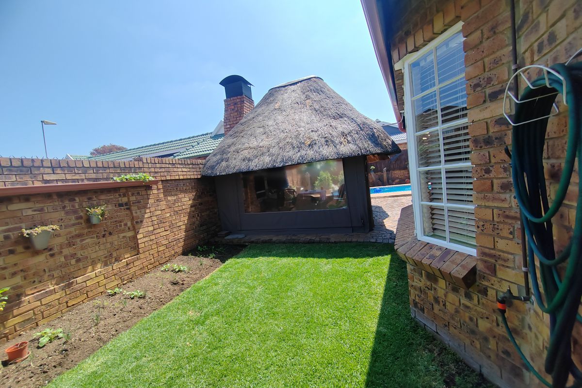 A well-maintained rear garden featuring a brick boundary wall, a manicured lawn, and a thatched lapa structure with a chimney.