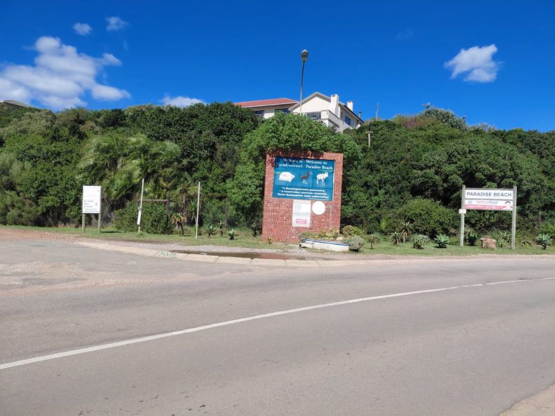 Street view of the Paradise Beach entrance featuring a brick signage wall, perimeter fencing, and surrounding coastal vegetation.