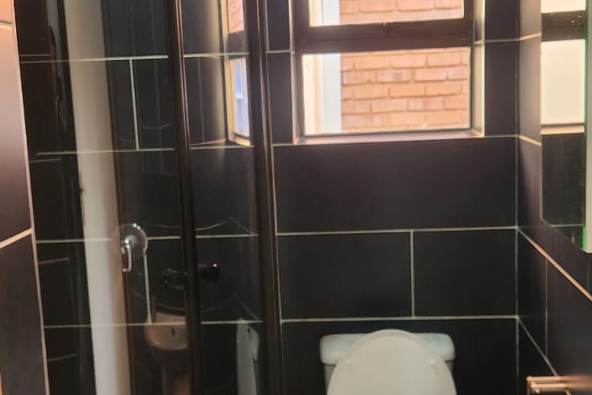 A compact bathroom featuring dark tiled walls, a corner shower enclosure, a pedestal basin, and a toilet, finished with a timber ceiling.