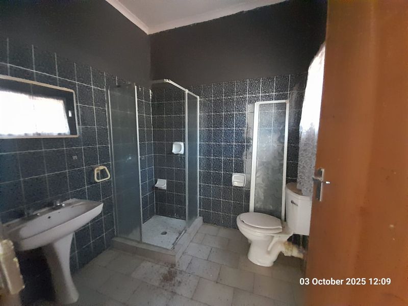 A dated bathroom featuring a pedestal basin, a shower enclosure with a missing panel, and a standard toilet, finished with dark wall tiling.