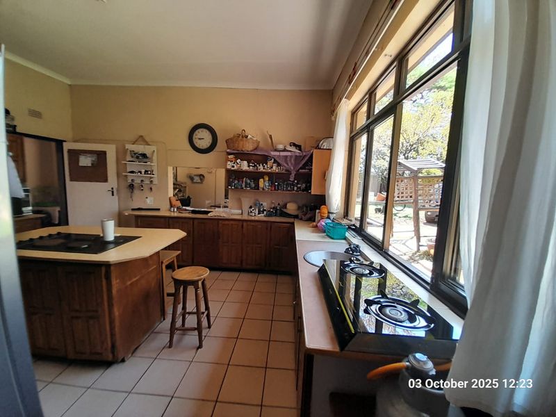 A functional kitchen featuring wooden cabinetry, a central island with a built-in hob, tiled flooring, and large windows overlooking the garden.