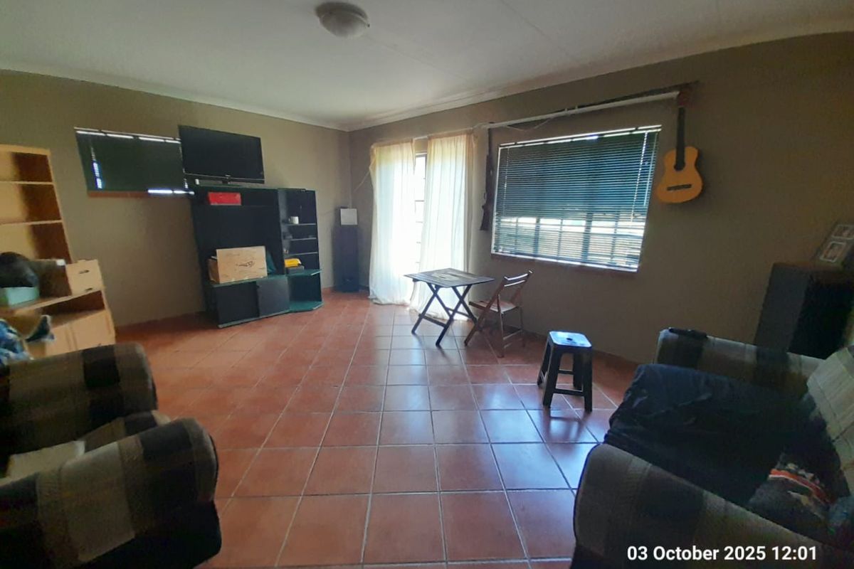 A spacious living room featuring terracotta floor tiles, large windows with blinds, and a sliding door providing access to the exterior.