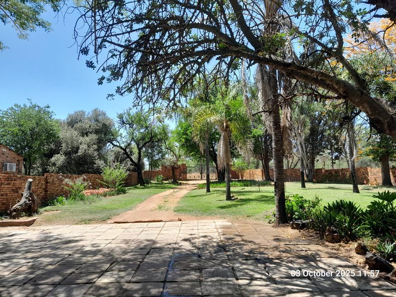 A spacious farm garden featuring a paved patio area, mature trees, established lawn, and a brick boundary wall with a gated entrance.
