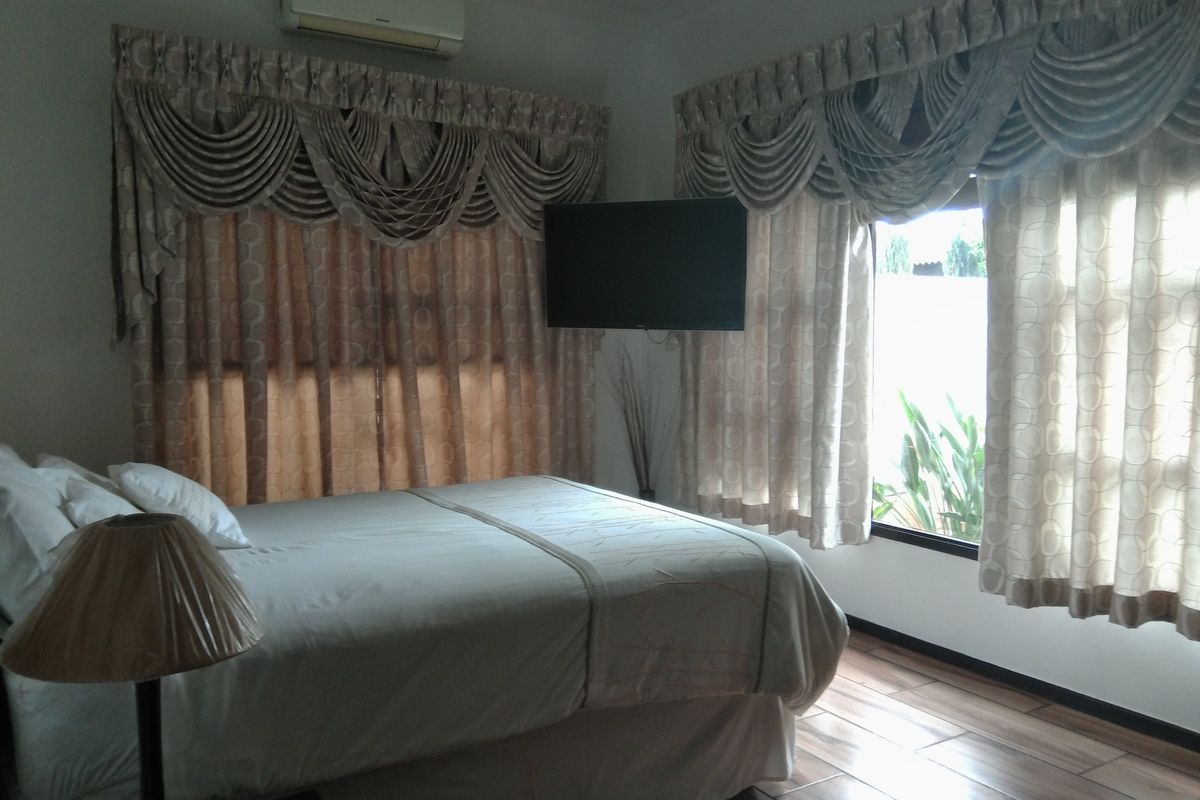 A bedroom featuring wood-look tile flooring, a wall-mounted air conditioning unit, and large windows with decorative valance curtains.