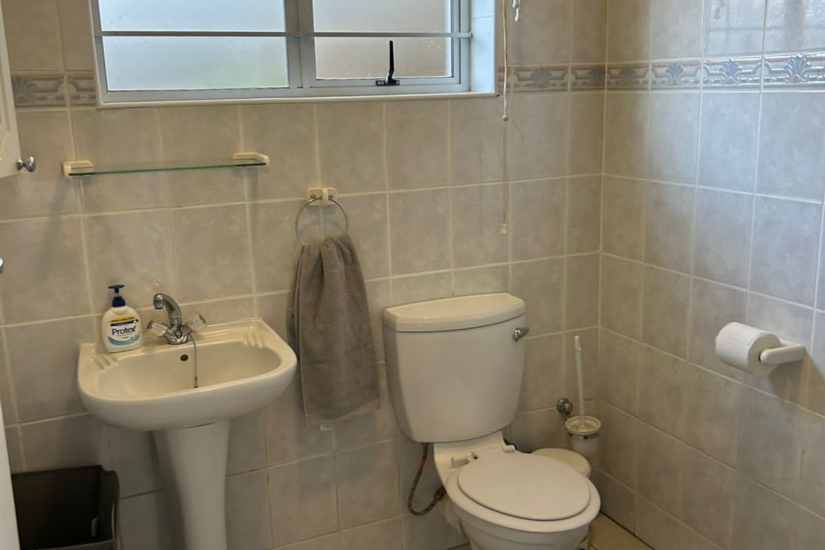 A functional bathroom featuring a pedestal basin, standard toilet, and floor-to-ceiling wall tiling with a window for natural light and ventilation.