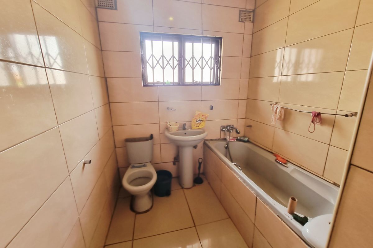 A dated bathroom featuring full-height wall tiling, a standard bathtub, pedestal basin, and toilet with security bars on the window.