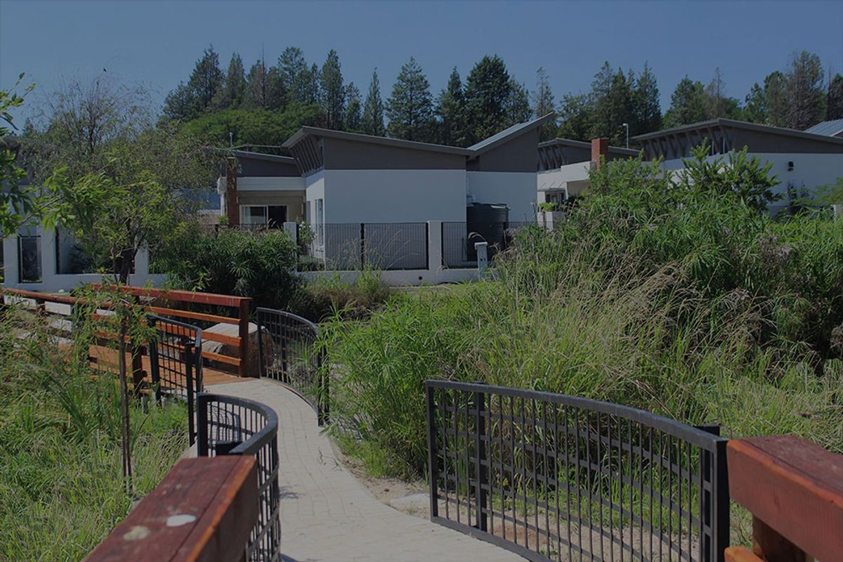 A scenic view of the townhouse complex featuring a paved walkway, a small bridge, and landscaped garden areas with mature trees.