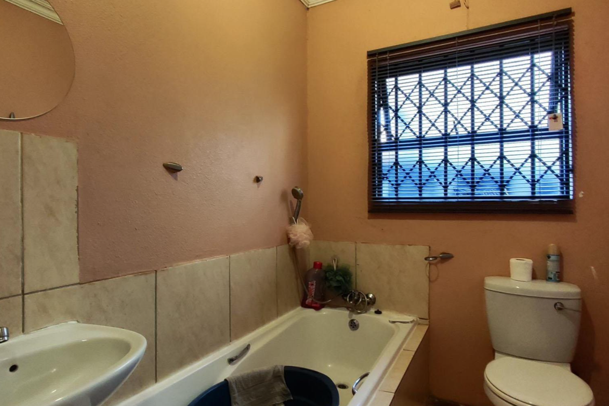 A dated bathroom featuring a standard bathtub, pedestal basin, and toilet, with a security-grille window and neutral wall tones.