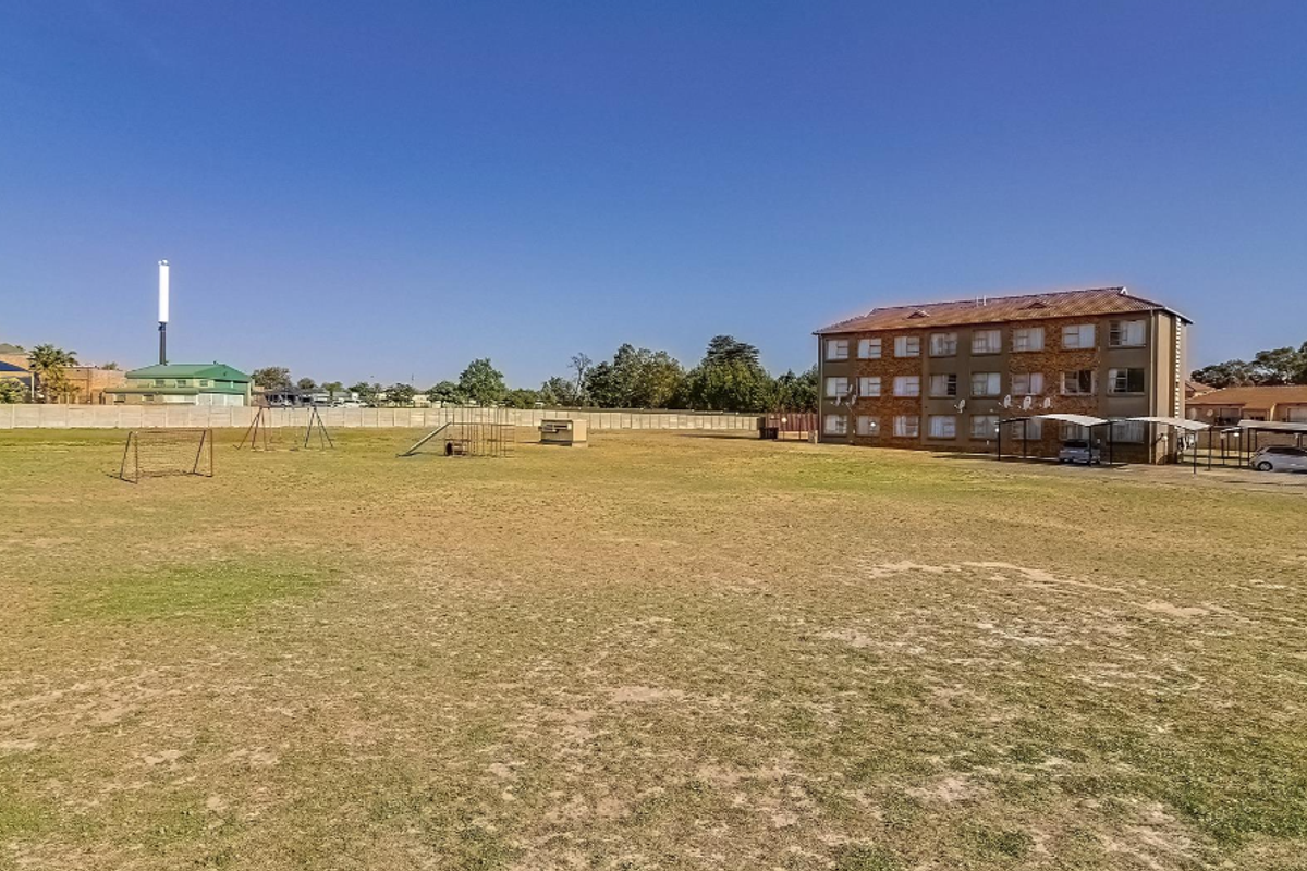 A wide-angle view of the apartment complex grounds featuring an open grass field, communal play area, and covered parking bays.