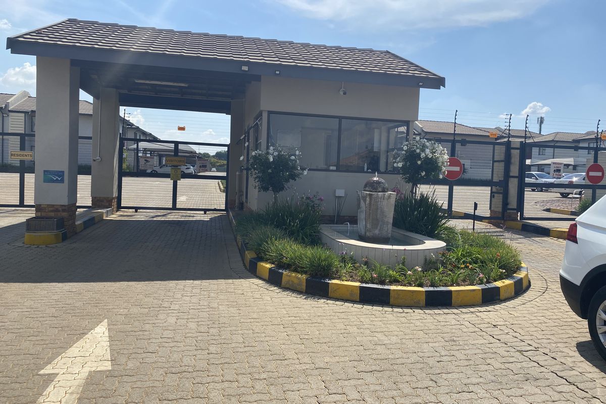 The secure entrance gatehouse of the townhouse complex, featuring a paved driveway, security boom gates, electric fencing, and a landscaped water feature.