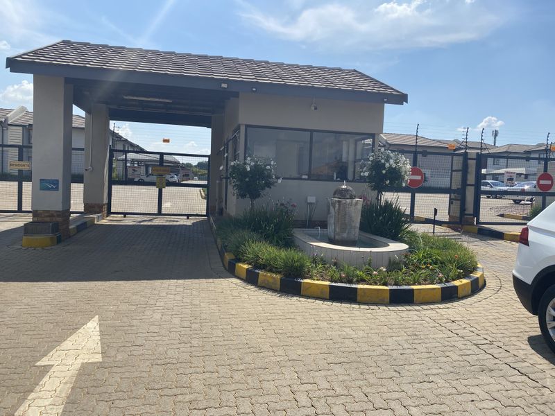 The secure entrance gatehouse of the townhouse complex, featuring a paved driveway, security boom gates, electric fencing, and a landscaped water feature.