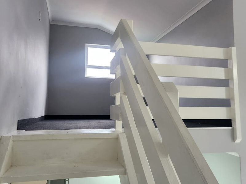 A loft-style bedroom area featuring a white wooden staircase, carpeted flooring, and a window providing natural light.