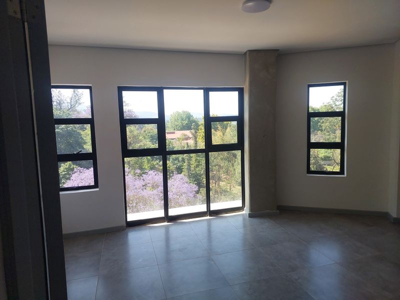 A bright, empty bedroom featuring large black-framed windows that offer scenic views of the surrounding trees and greenery.