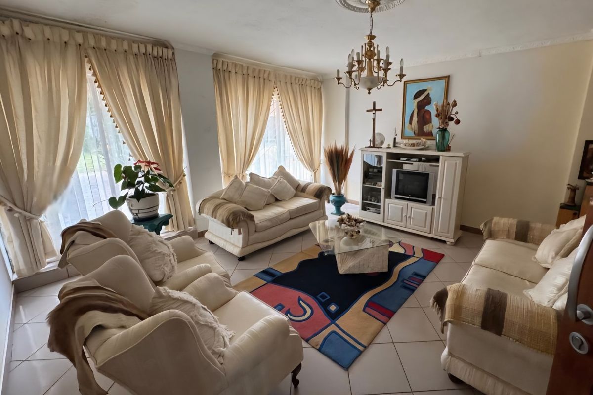A traditional living room featuring tiled flooring, decorative ceiling rose with chandelier, and large windows with floor-to-ceiling curtains.