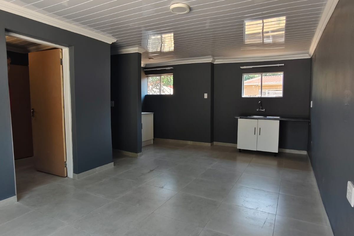An open-plan living area featuring grey tiled flooring, a modern PVC ceiling, and a compact kitchenette with base cabinetry and a sink.