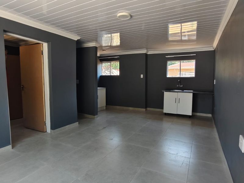 An open-plan living area featuring grey tiled flooring, a modern PVC ceiling, and a compact kitchenette with base cabinetry and a sink.