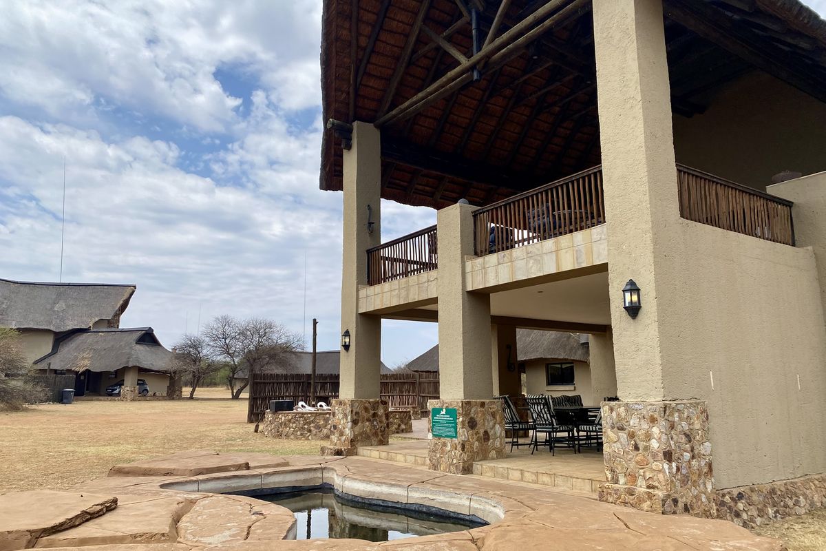 A two-story thatched-roof residence featuring a stone-clad pool area, covered patio, and balcony, set within a spacious natural landscape.