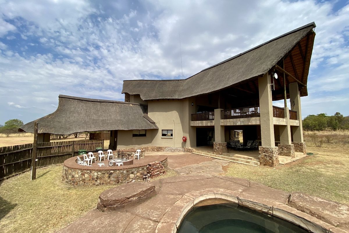 A spacious double-story home featuring a traditional thatched roof, a stone-clad fire pit area, and a natural rock-style swimming pool in a rural setting.