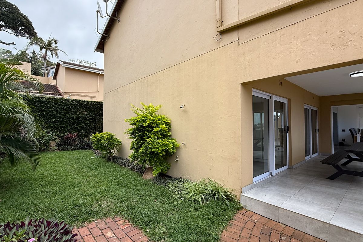 A rear view of the apartment featuring a covered patio with tiled flooring, leading out to a private garden area with established landscaping.