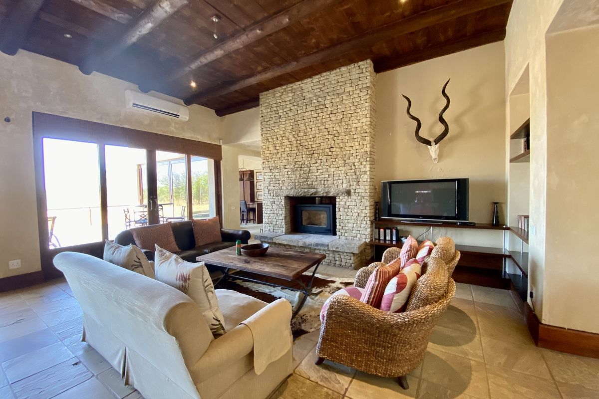 Spacious living room featuring a prominent stone fireplace, exposed timber beam ceiling, tiled flooring, and large sliding glass doors leading to the outdoor area.