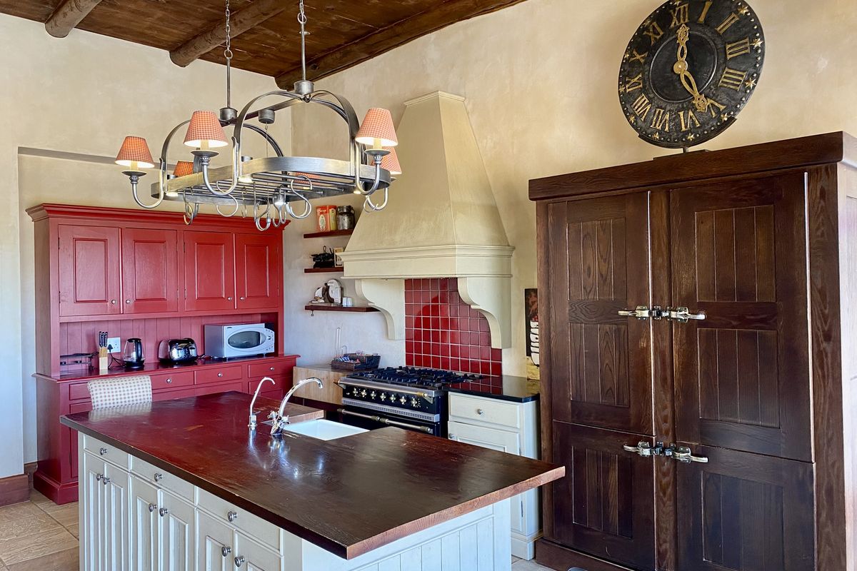 A rustic-style kitchen featuring a large central island with a wooden countertop, red cabinetry, a built-in range with a tiled backsplash, and exposed timber ceiling beams.