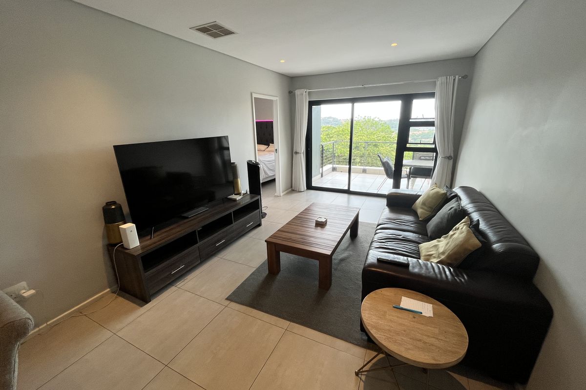 A modern living room featuring large format floor tiles, recessed downlighting, and sliding glass doors that open onto a private balcony with scenic views.