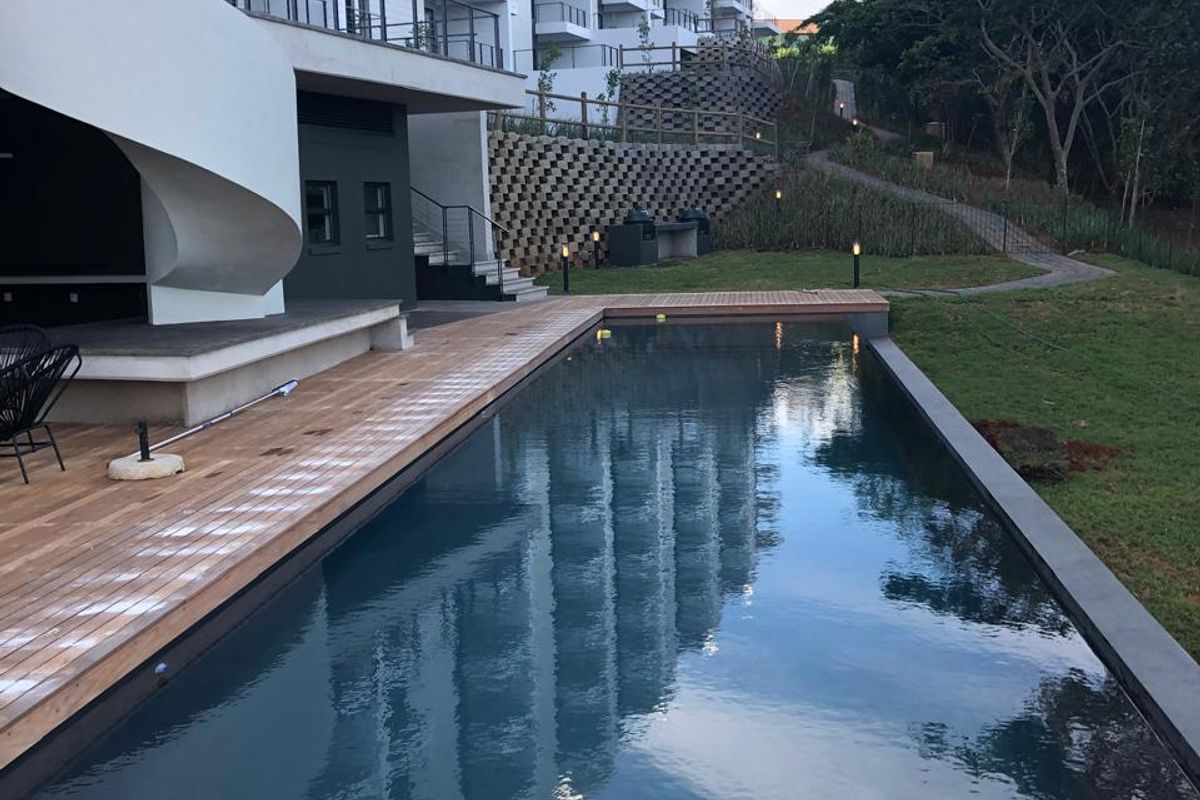 A modern swimming pool area featuring a timber deck, landscaped garden, and views of the apartment building facade.