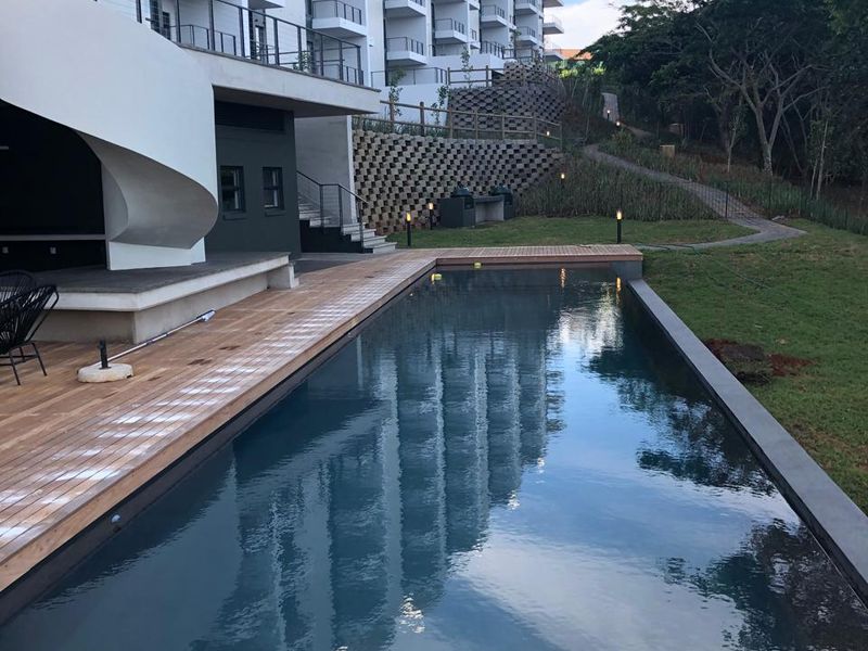 A modern swimming pool area featuring a timber deck, landscaped garden, and views of the apartment building facade.