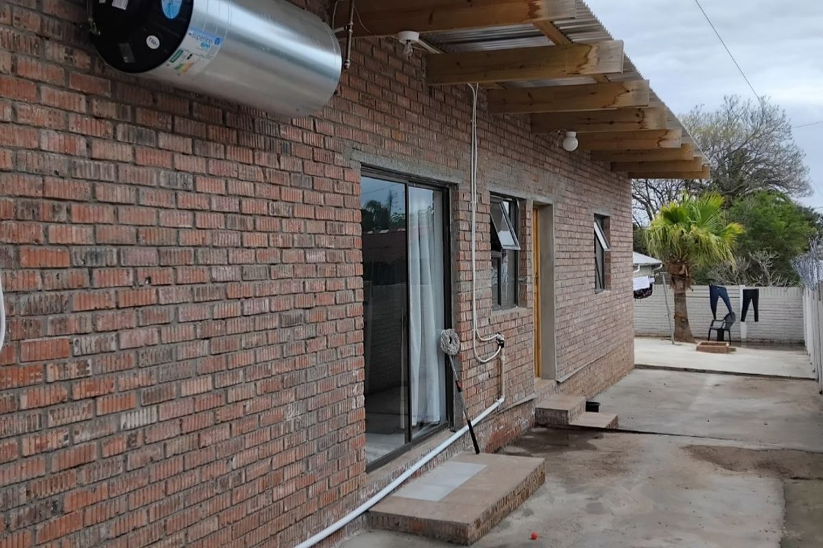 Rear exterior view of a brick house featuring a corrugated metal roof, exposed timber rafters, and an externally mounted electric water heater.