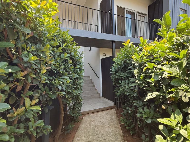 A well-maintained apartment entrance featuring a paved walkway, concrete staircase, and modern metal balustrades, framed by established garden hedging.