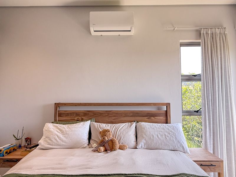 A well-maintained bedroom featuring a wall-mounted split-system air conditioner, large window with natural light, and built-in wardrobe storage.