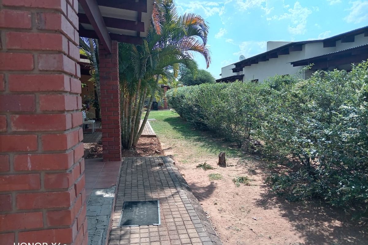 A view of the exterior rear area featuring a covered patio, brick pillars, paved walkway, and a garden with established hedges.